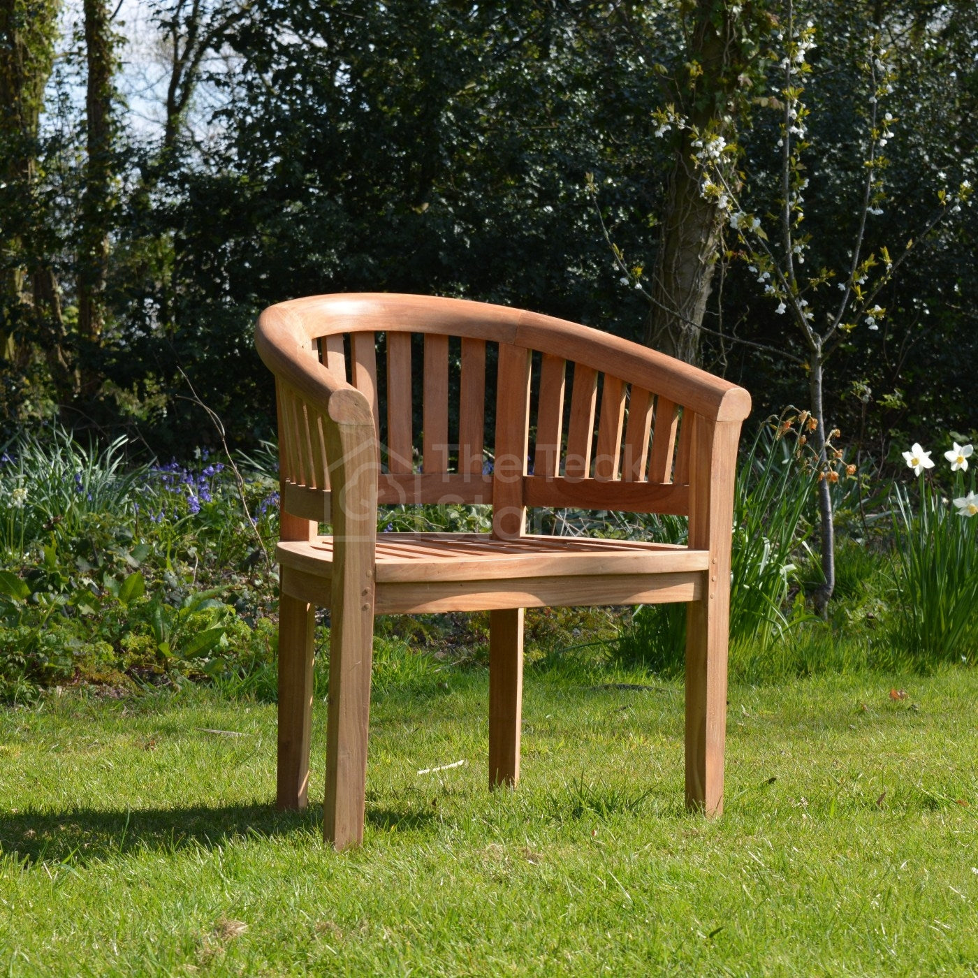 Angled side view of the Lulworth Teak Banana Chair placed on grass, highlighting its rounded arms and slatted back.